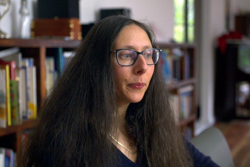 A woman sitting indoors looks off to the side with a worried expression on her face. She has long dark hair and glasses.