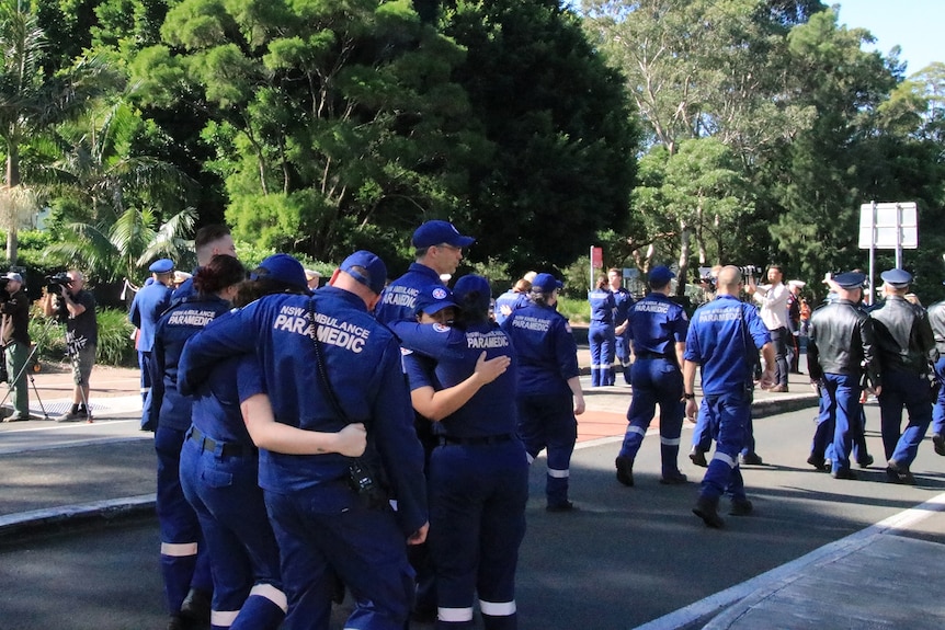 A group of paramedics wearing blue uniforms hug on a road.