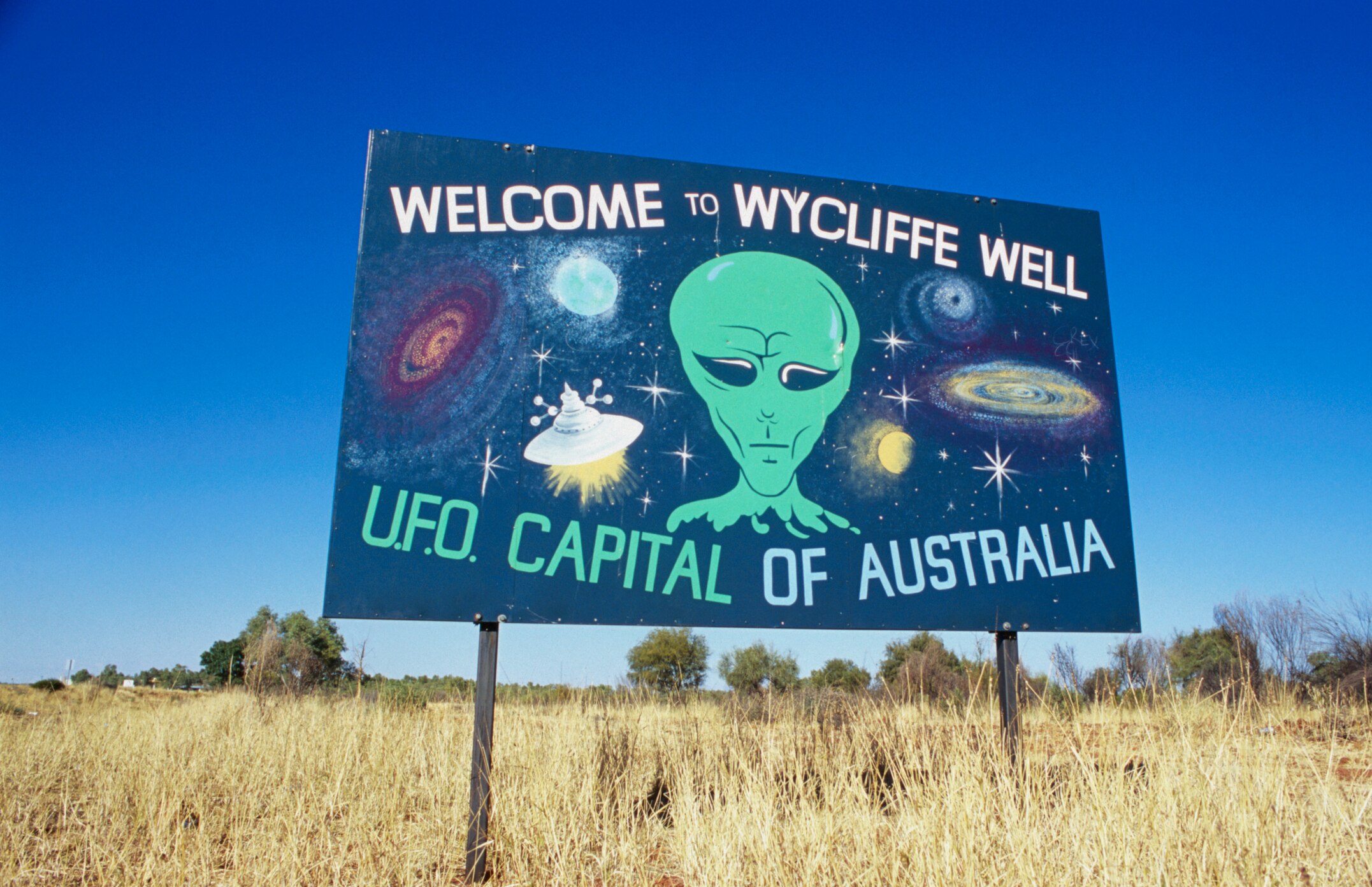 A sign in the outback welcoming visitors to Wycliffe Well, with a painting of a green alien.