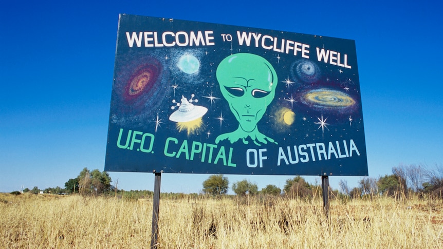A sign in the outback welcoming visitors to Wycliffe Well, with a painting of a green alien.
