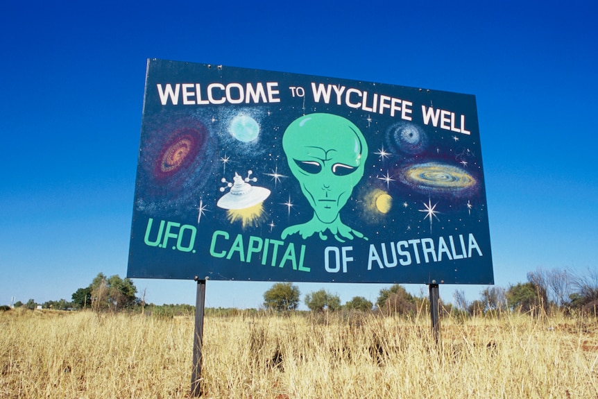 A sign in the outback welcoming visitors to Wycliffe Well, with a painting of a green alien.