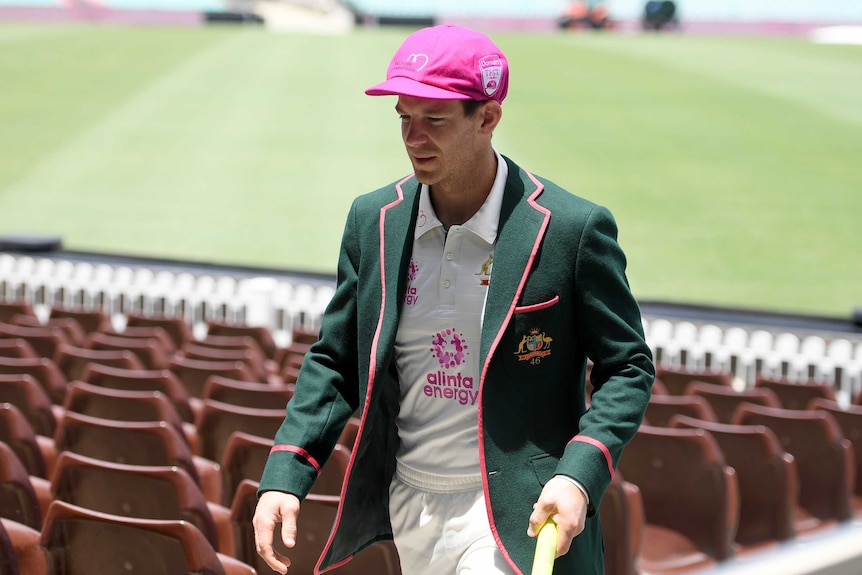 Wearing a green blazer, whites and a pink baggy cap, Tim Paine walks through up the SCG race past members seats