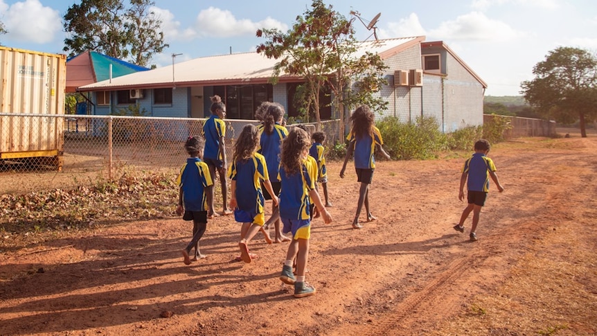 A group of primary aged kids in school uniforms walking on a red dirt road. 