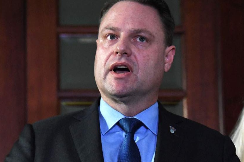 A man with dark hair, wearing a dark suit and flanked by a blonde, formally dressed woman speaks at a press conference.