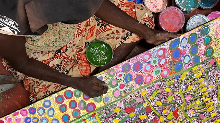 An overhead shot of an Indigenous woman painting a large floral atwork.