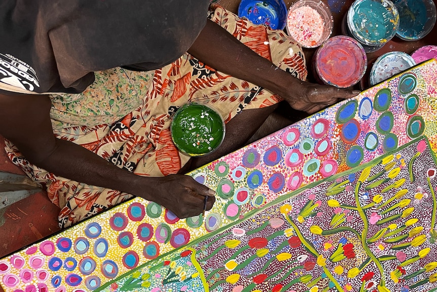 An overhead shot of an Indigenous woman painting a large floral atwork.