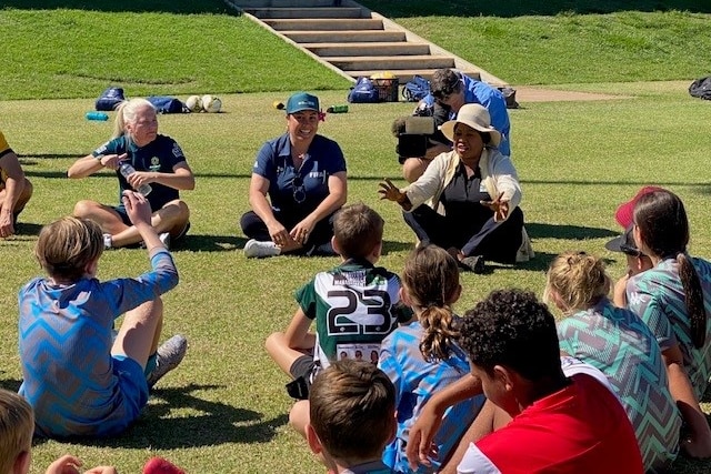 A FIFA football executive with a broad-brimmed hat on her head sits in a circle on the grass and talks to young footballers.