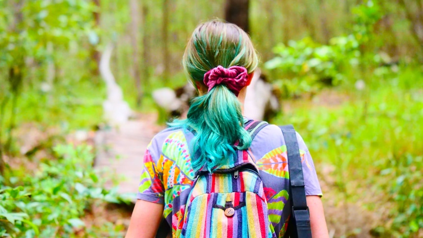 a woman with dyed green hair and a brightly coloured top and backpack hikes in bushland
