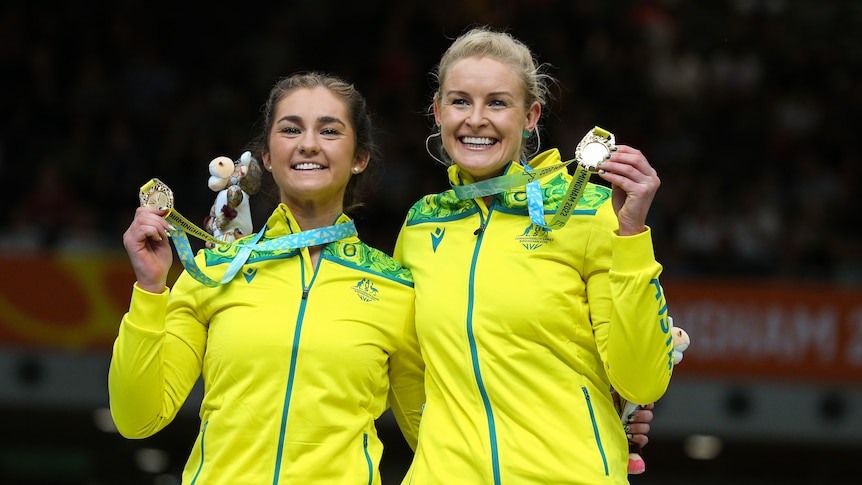 Two female athletes wearing yellow and green hold up gold medals