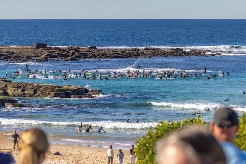 Blurred out backs of heads of onlookers viewing hundreds of people paddling out to sea on surfboards in the distance.