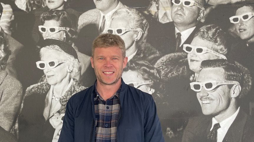 A man with short blonde hair and a blue shirt stands in front of an old cinematic image of men and women watching a 3D movie