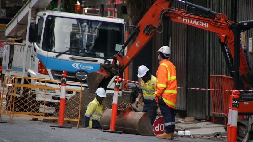 Three construction workers stand between machinery at a worksite.