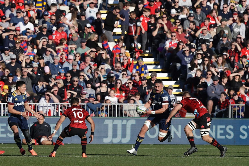 A rugby player runs at opponents while carrying the ball in a stadium packed with people wearing team colours.