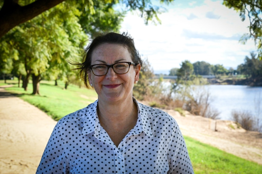 A close up of a smiling woman in a white collared shirt with navy polka dots and black glasses.