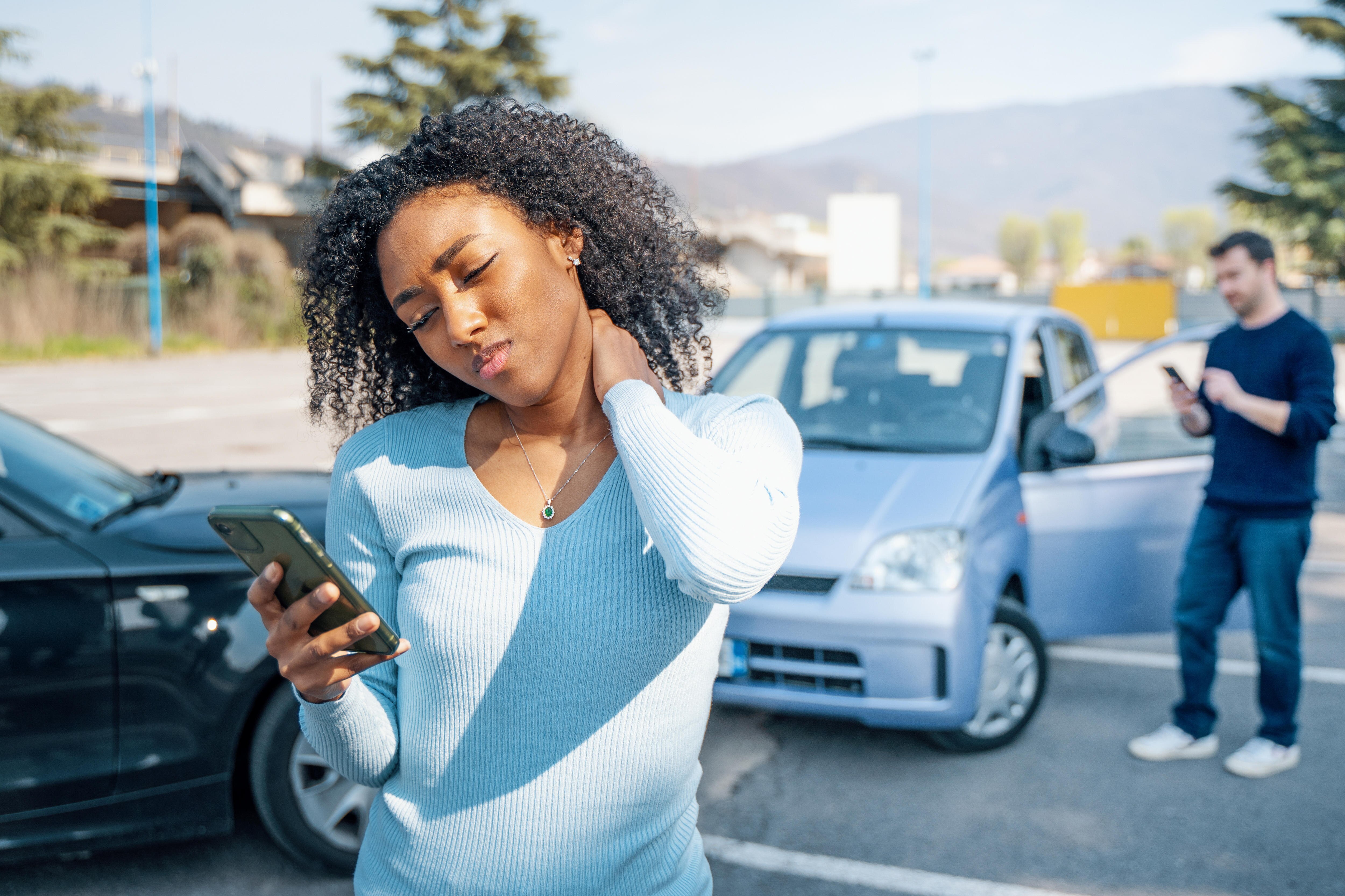 A stressed woman looks at her phone in front of a minor car crash.