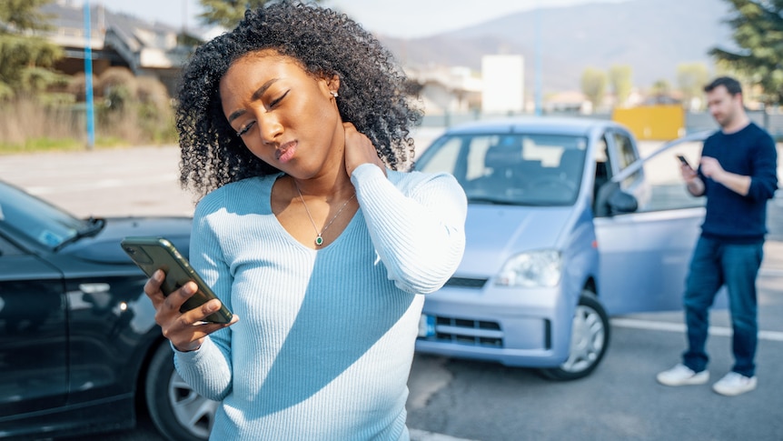 A stressed woman looks at her phone in front of a minor car crash.