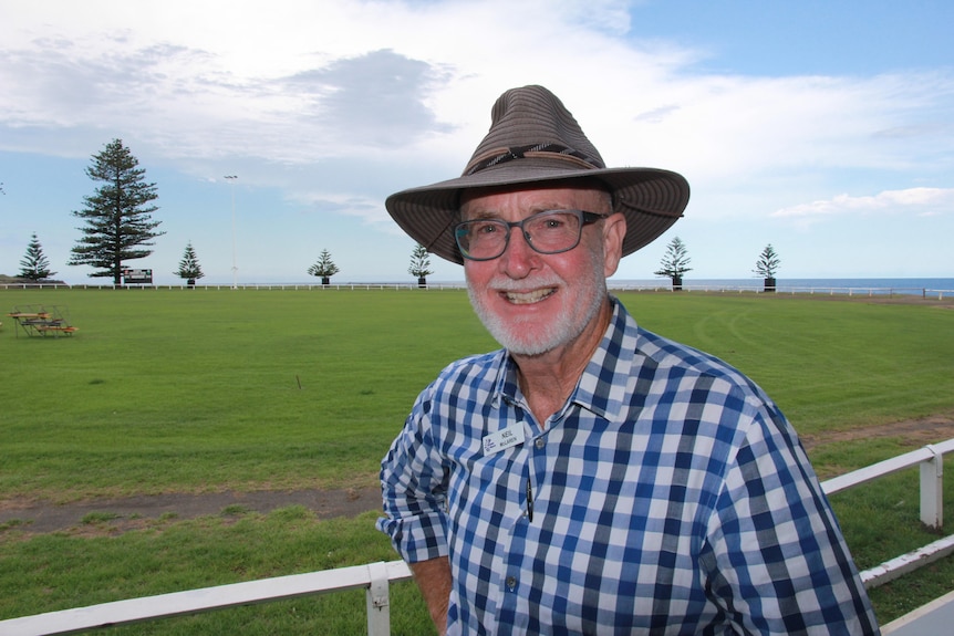 A smiling, middle-aged man in a hat and chequered shirt standing in front of a lush field.