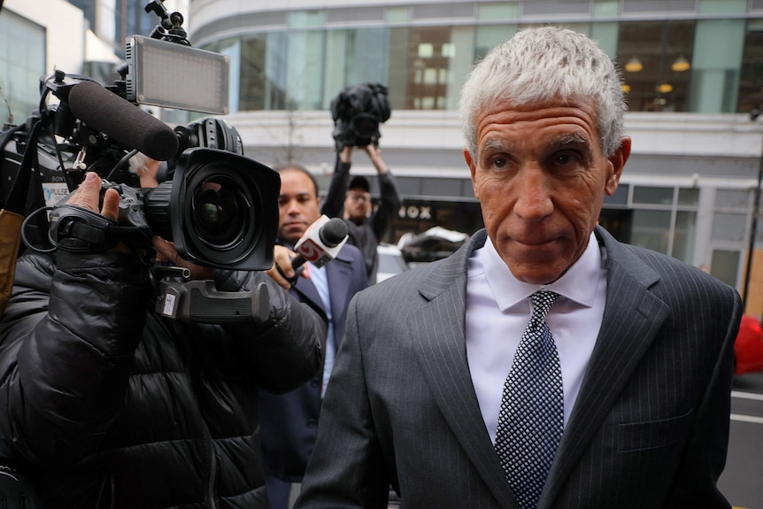 A middle-aged man with grey hair arrives outside court in a grey suit. Media surrounds him.