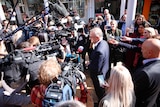 A man surrounded by a ring of journalists holding cameras and microphones
