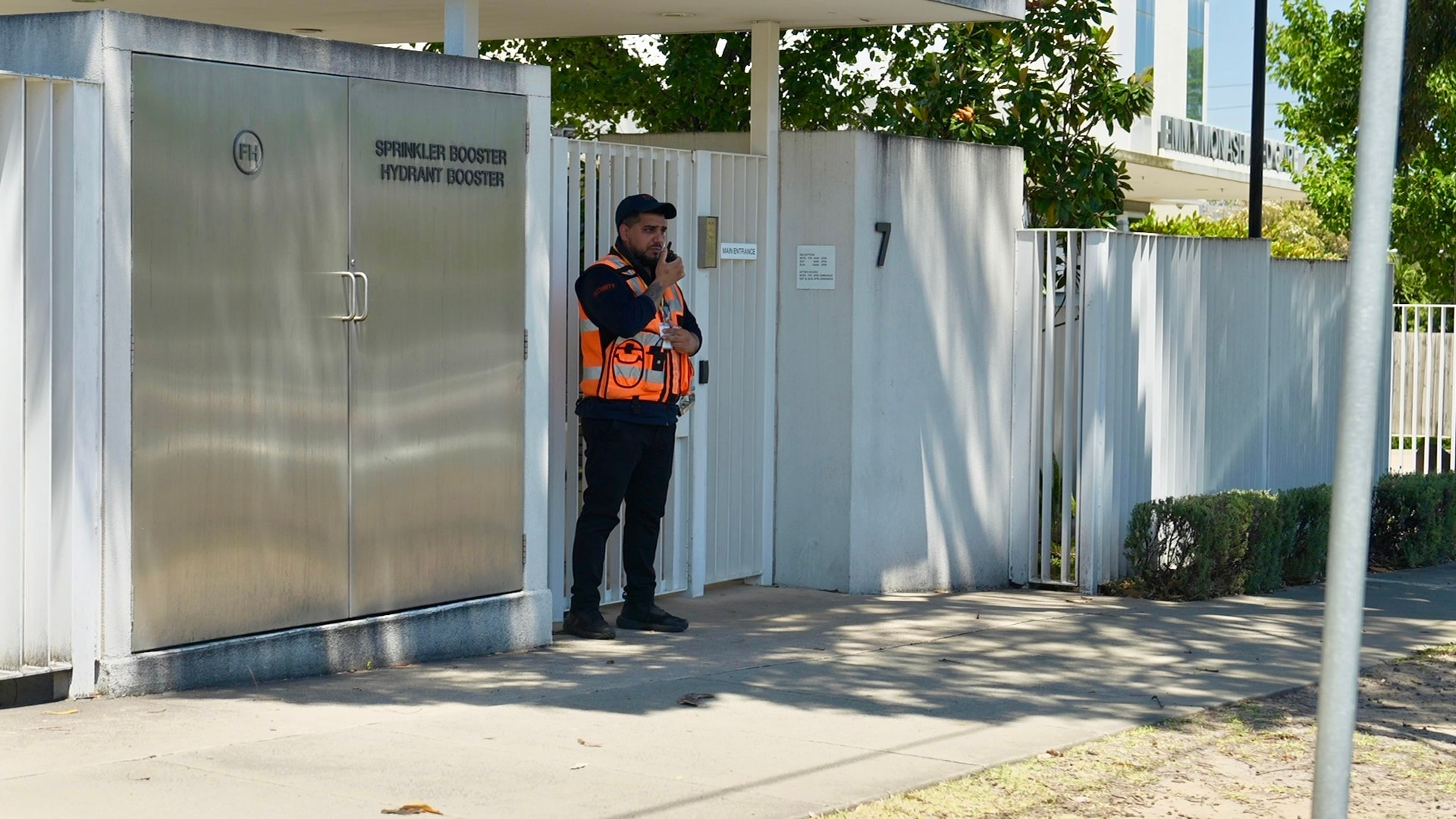 Um homem de boné preto, colete laranja de alta visibilidade e calça preta fala em um walkie-talkie próximo a uma cerca branca.