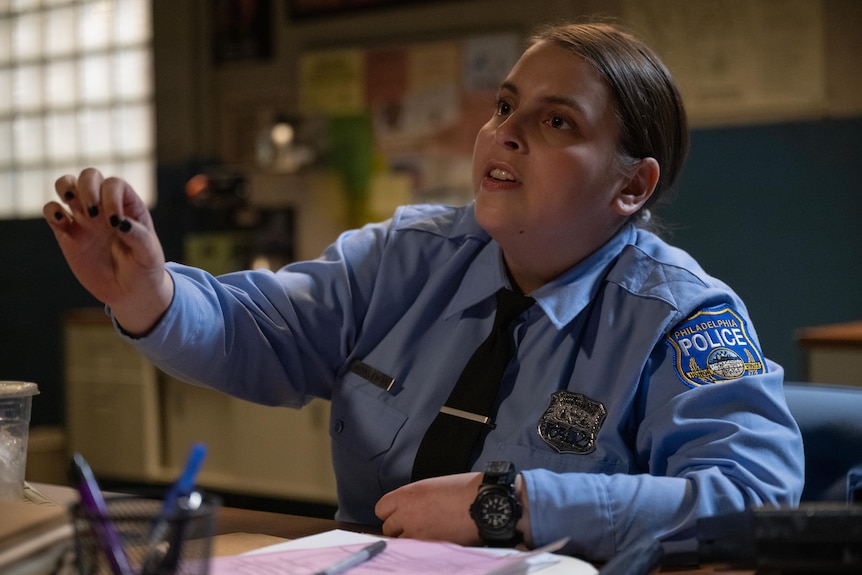 A film still of Beanie Feldstein, wearing a police uniform, and sitting at a desk. She has one arm outstretched.