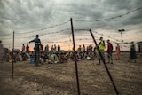 A man directs people to sit in different queues on dry ground behind a barbed wire fence