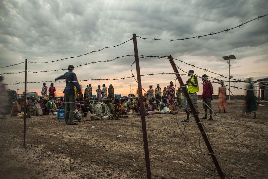 A man directs people to sit in different queues on dry ground behind a barbed wire fence