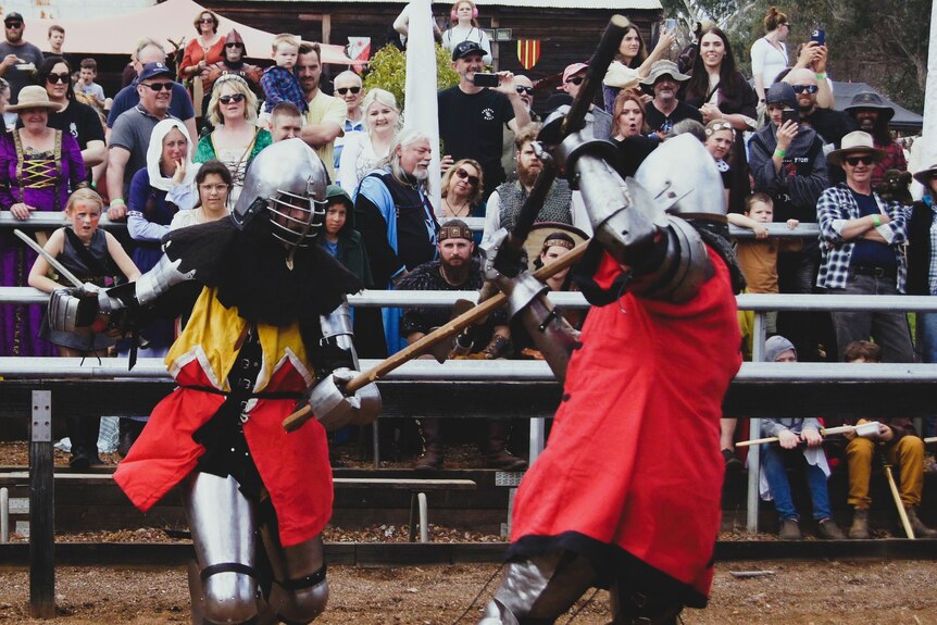 Two people in Medieval armour, armed with weapons, fight as a crowd watches on