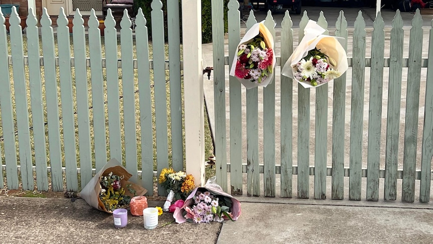 Bunches of flowers on the ground by a picket fence and a few bouquets hanging from the fence. 