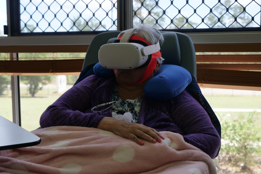 A grey haired woman sits in a chair with white virtual reality goggles over her head and a pink blanket over her lap.