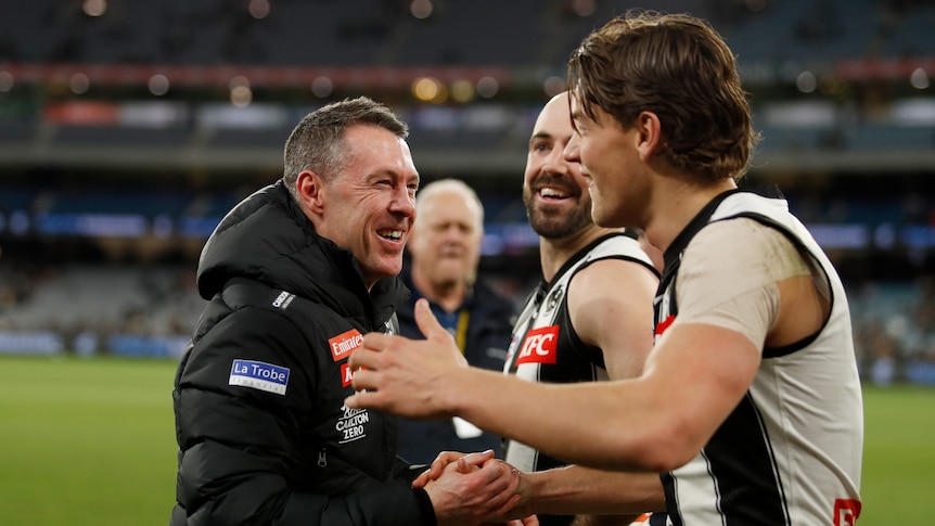 An AFL coach wears a beaming smile as he clasps hands with one of his players after an important victory.