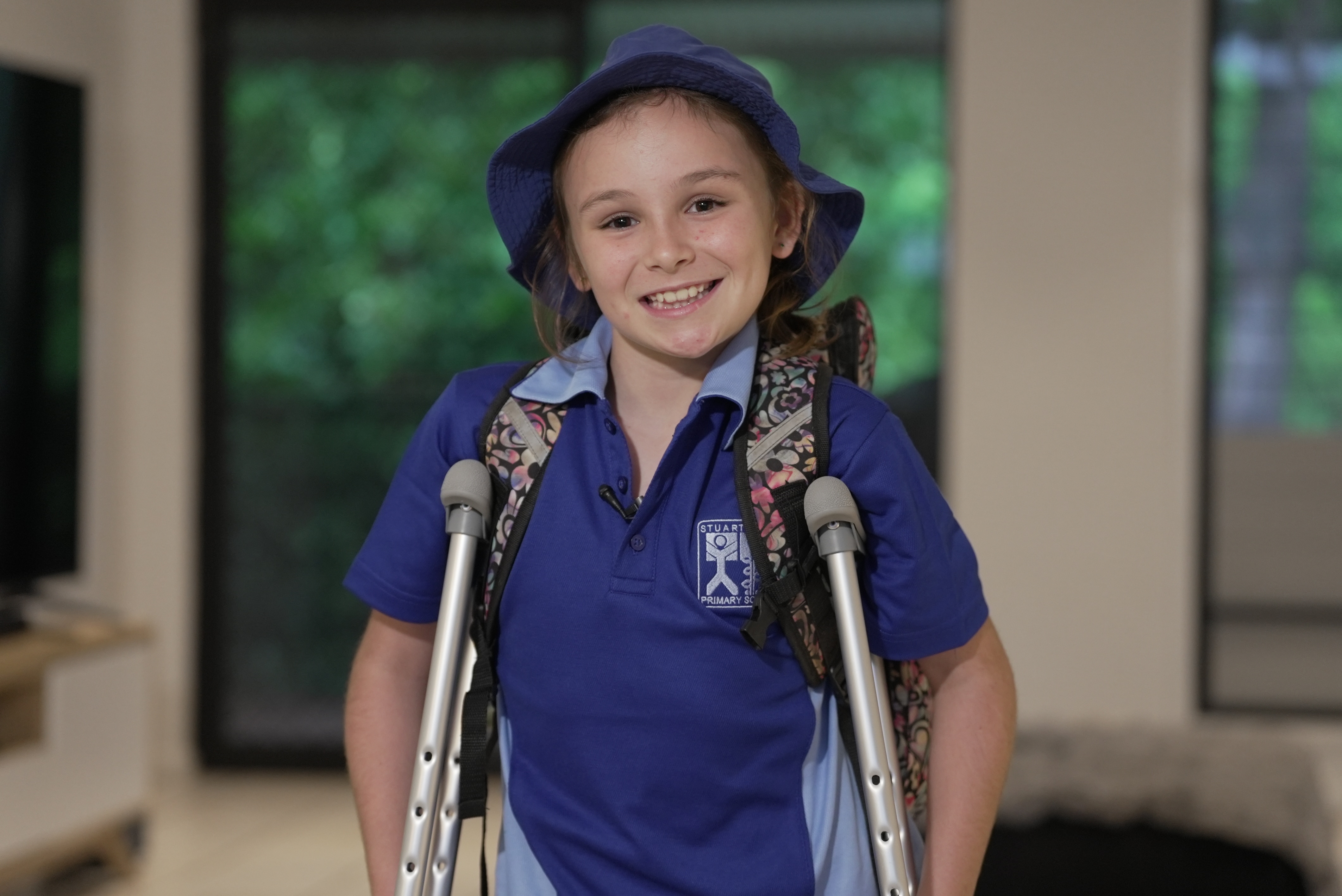 Uma menina vestida com seu uniforme azul da escola primária e segurando muletas enquanto sorri para a câmera.