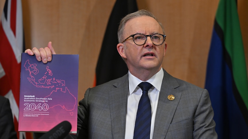 Anthony Albanese in a grey suit and tie holds up a purple booklet in front of three flags