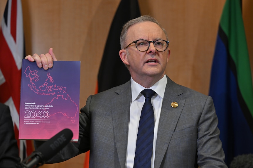 Anthony Albanese in a grey suit and tie holds up a purple booklet in front of three flags