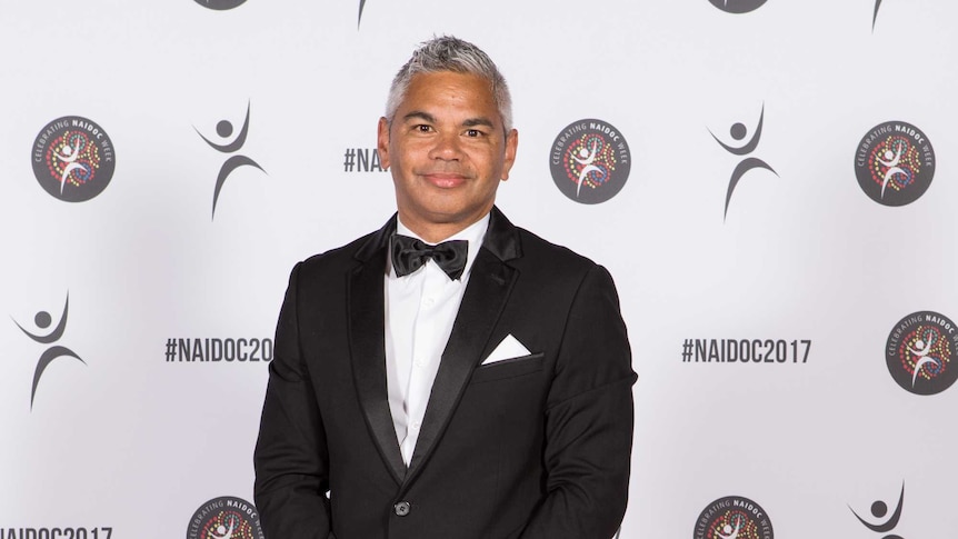 A man with silver hair stands in front of a white and black media wall wearing a dinner suite and bow-tie.