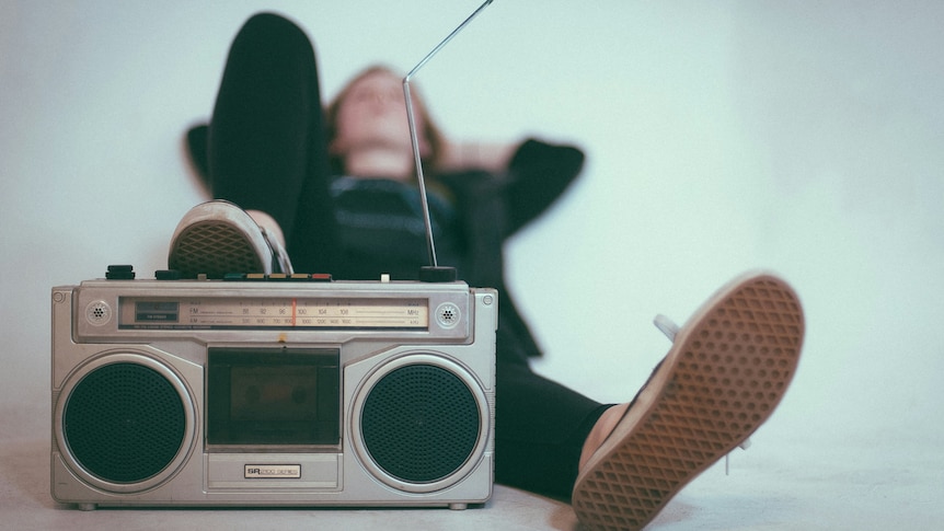 An 80's boom box radio sits at the front of the image. A person lies on their back behind it with one foot on the stereo.