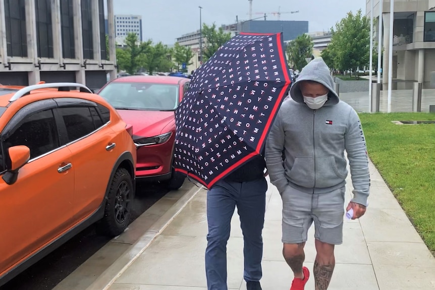 Two men walk outside the courts. One man's face is covered by an umbrella.