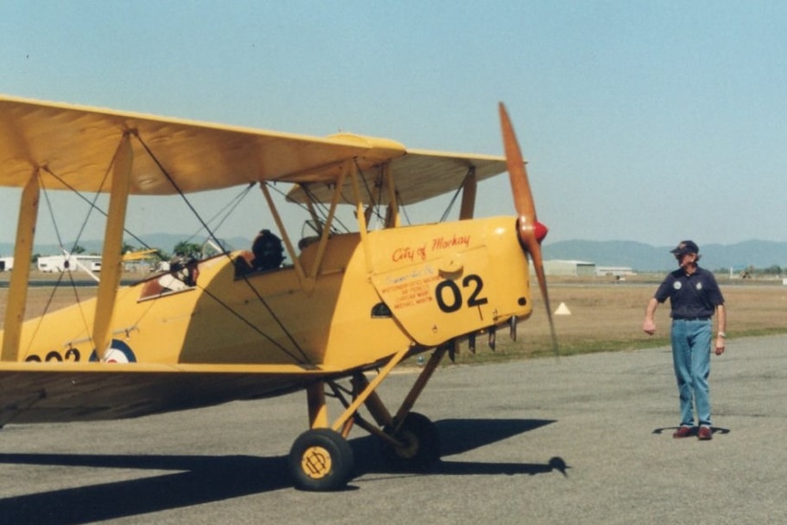 an old yellow plane with an open cockpit sits on a runway and a crew member is standing alongside it