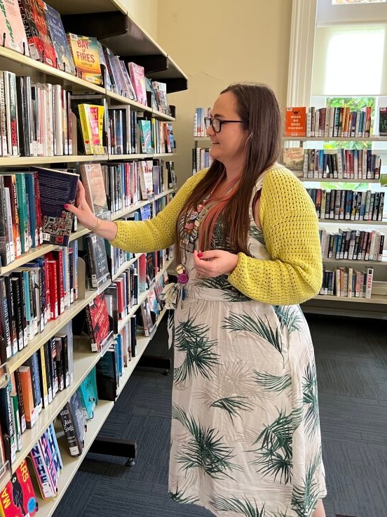 A woman with long brown hair, wearing a green and white dress and a yellow cardigan pulls a book from a shelf at a library.