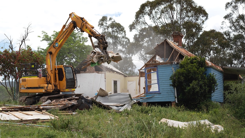 A blue wooden home with an excavator destroying ripping down walls. 