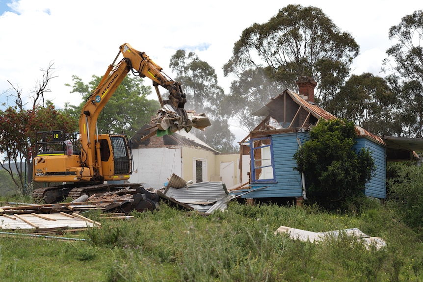 A blue wooden home with an excavator destroying ripping down walls. 