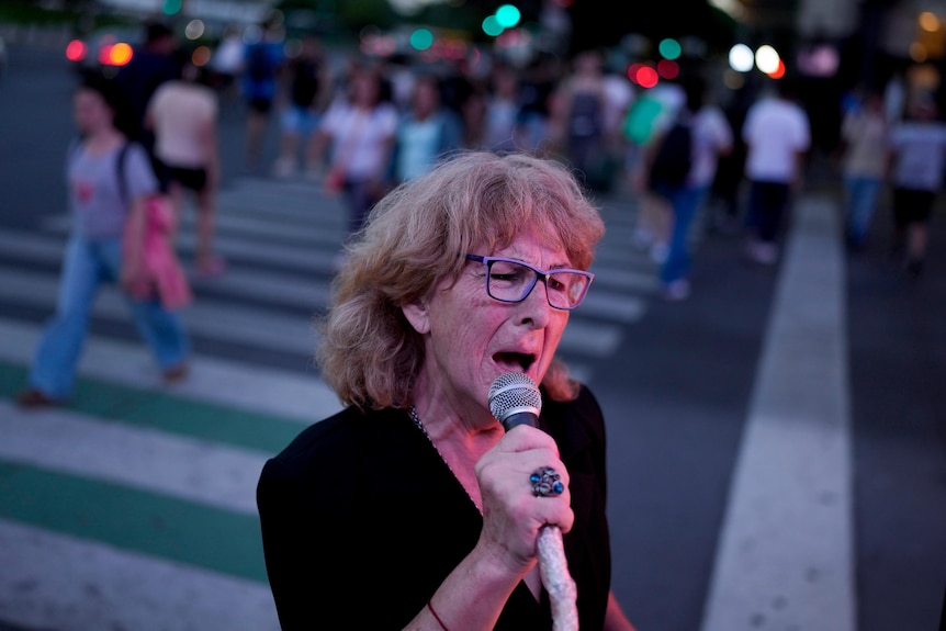 An older woman holding a microphone sings in front of a crowd of people on a crossing behind her.