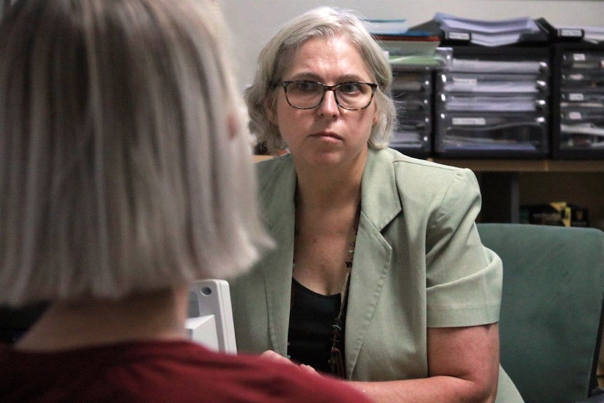 A mid shot of a female doctor with grey hair and glasses sitting in an office with a patient's head in the foreground.