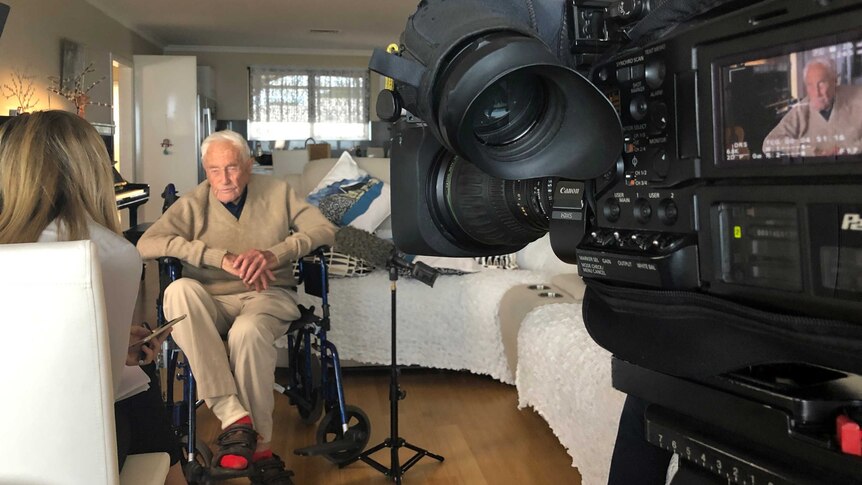 A wide shot showing David Goodall being interviewed in his living room by Charlotte Hamlyn with a TV camera to the right.