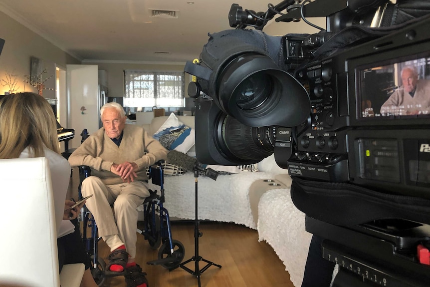 A wide shot showing David Goodall being interviewed in his living room by Charlotte Hamlyn with a TV camera to the right.