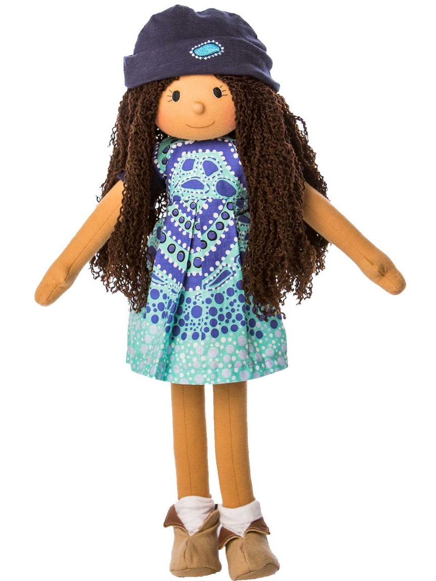 A doll with brown skin, long dark hair, a beanie and a light blue dress with an Indigenous design.