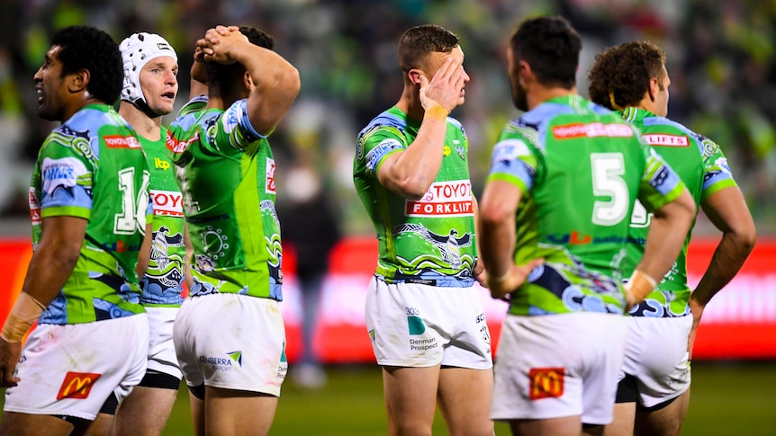 A group of dejected NRL teammates stand with hands on heads as they wait for the other team to kick the conversion after a try.