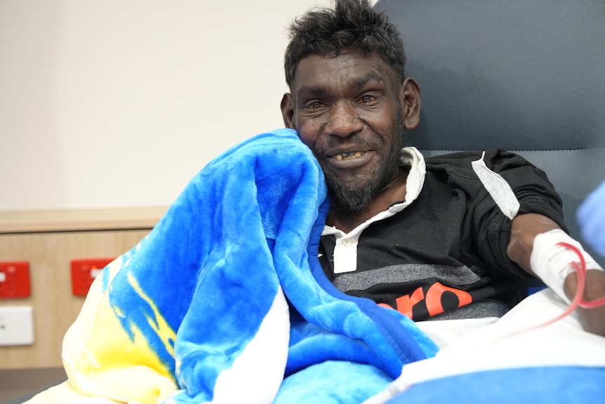 A photo showing an indigenous man sitting back in a leather hospital chair wrapped in blue blanket.