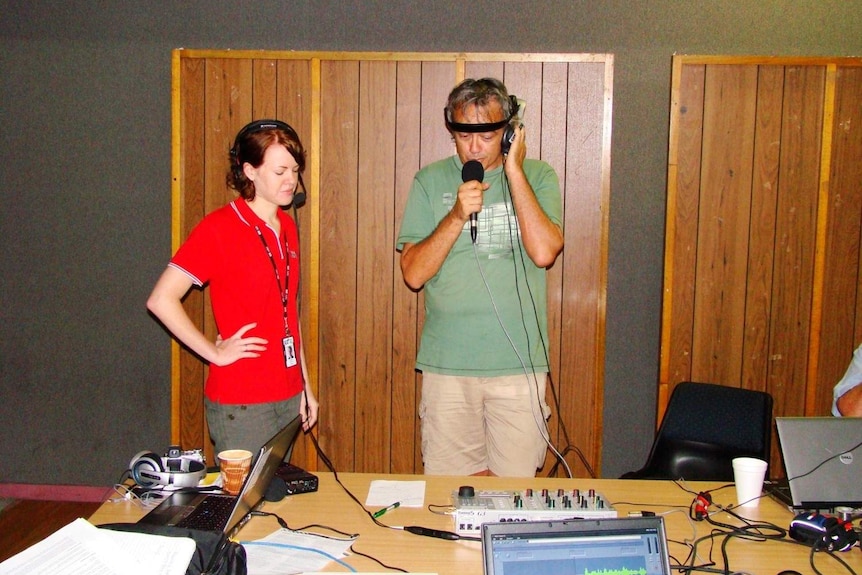 Dinnen and Denyer wearing headphones and standing at desk with mobile radio equipment inside a building.