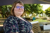 A woman in glasses sits on a bench in a park.